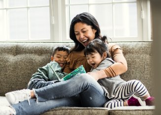 duas crianças com mulher em sofá olhando livro e rindo