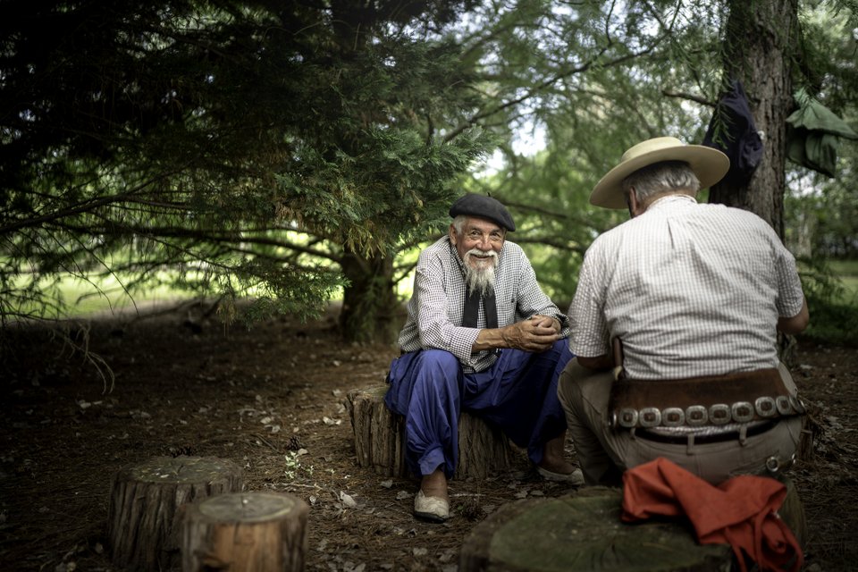 dois senhores sentados em troncos de árvore com roupas e acessórios de trajes típicos gaúcho