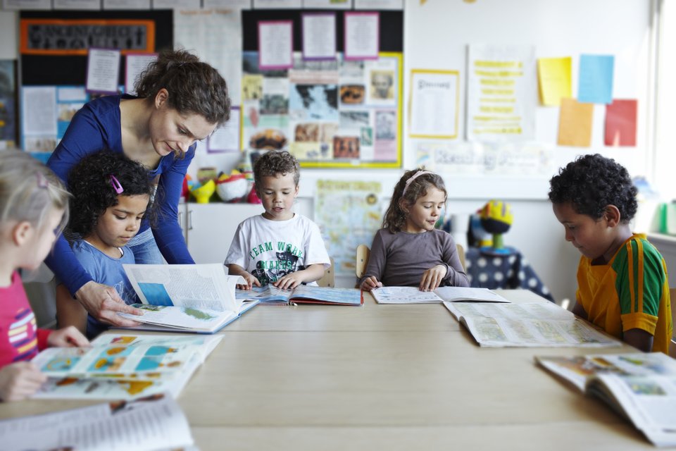 adulto auxiliando as cinco crianças a olharem o livro em uma sala de aula
