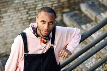 homem negro com fone de ouvido colorido no pescoço, blusa de manga comprida rosa e jardineira jeans