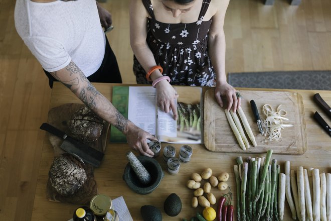 mãos e braços de um homem e uma mulher em cima de uma mesa com vários alimentos para serem preparados, além de um livro de receitas