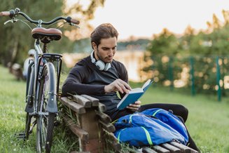 homem lendo livro no parque sentado em banco com uma bicicleta encostada no banco