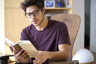 homem de óculos sentado em poltrona com livro aberto nas mãos