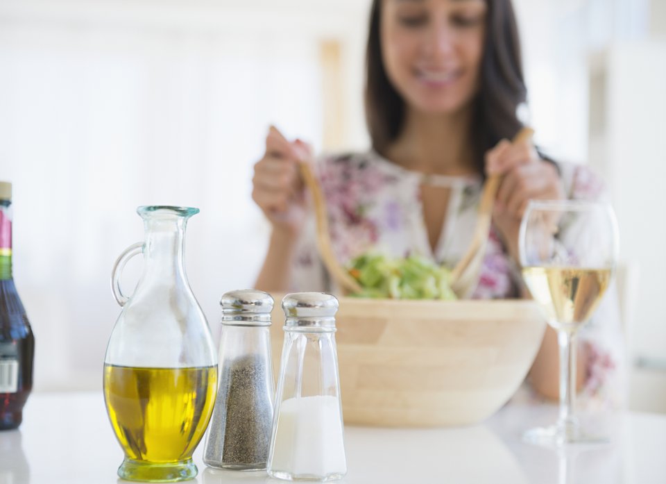 galheteiro saleiro e pimenteiro a frente e ao fundo da mesa, uma mulher desfocada mexendo uma salada