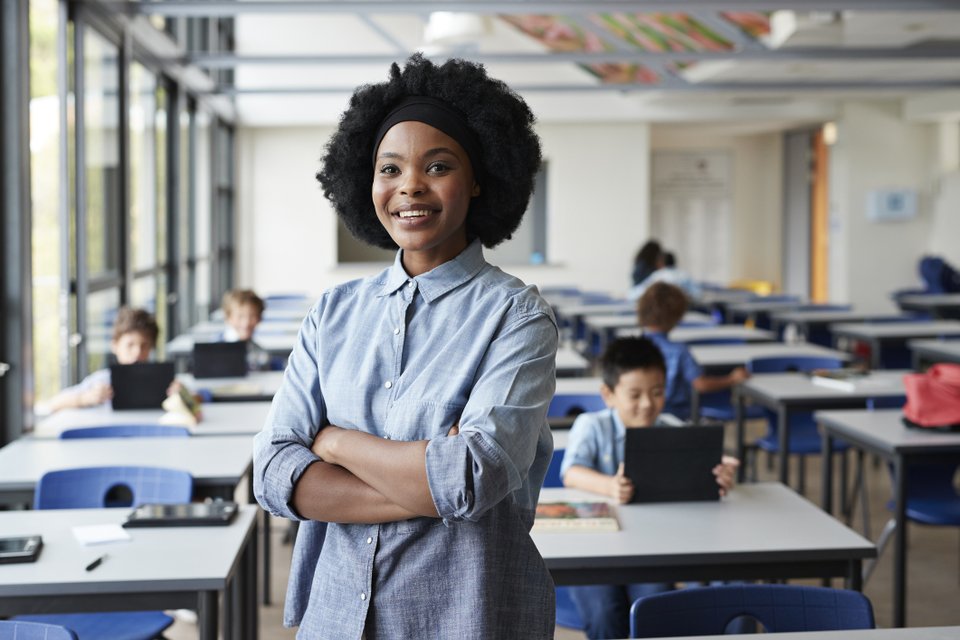 mulher negra com camisa azul e ao fundo uma sala de aula com alguns alunos