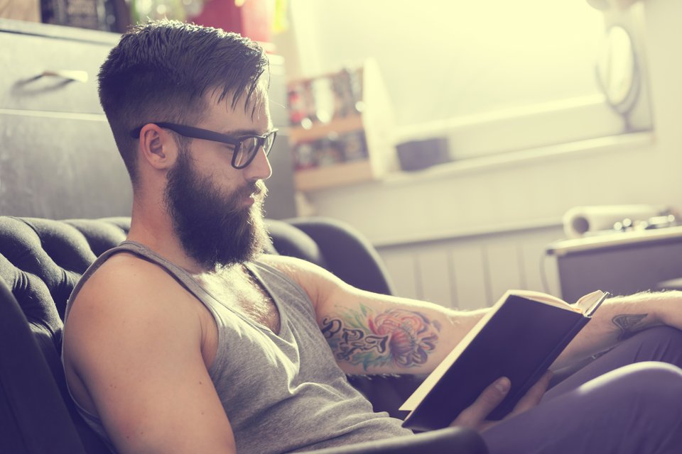 homem de barba, óculos e tatuagem no braço, lendo um livro sentado em um sofá