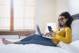 mulher deitada na cama com calça e blusa de manga amarela segurando notebook e celular