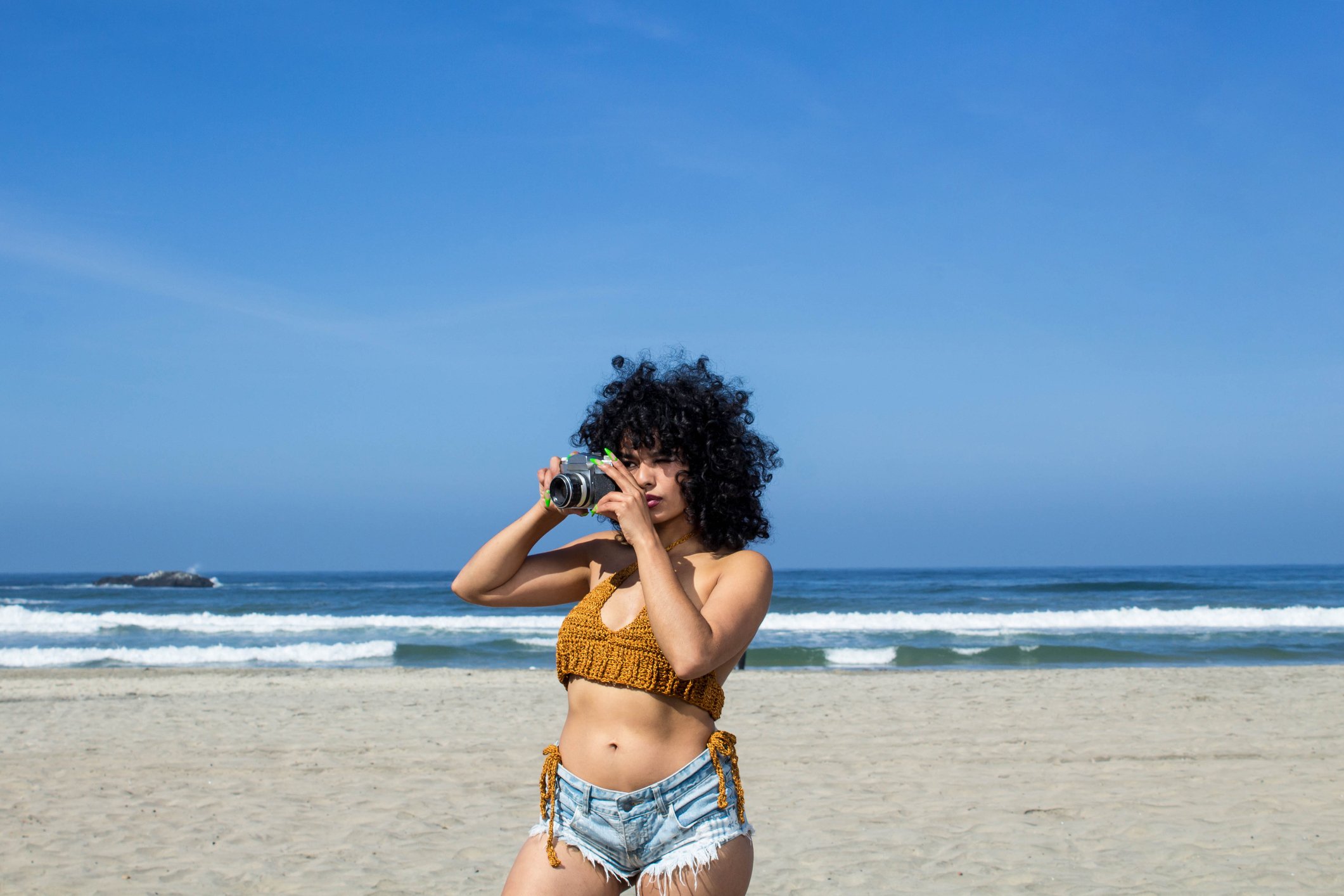 mulher com biquíni de crochê e short jeans na praia usando máquina fotográfica