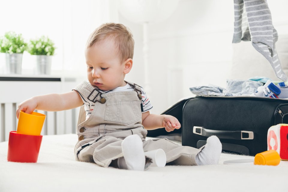 criança em cima de uma cama brincando e ao lado uma mala de viagem