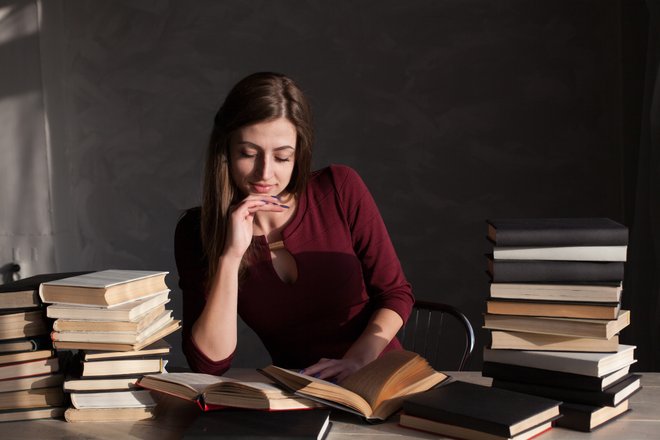 mulher com mão no queixo e outra mão segurando um livro num cenário com vários livros em cima da mesa