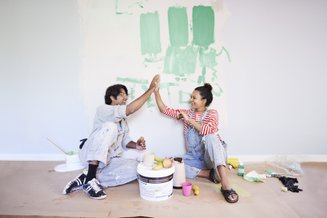 homem e mulher sentados no chão encostados numa parede branca com tinta verde. ao chão, latas de tinta