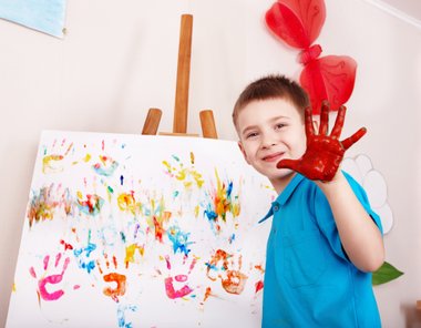 criança com mão com tinta e quadro cheio de mãos coloridas de tinta atrás em cima de um cavalete
