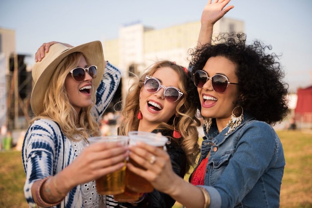 Três jovens sorridentes em festival ao ar livre com copos de cerveja na mão