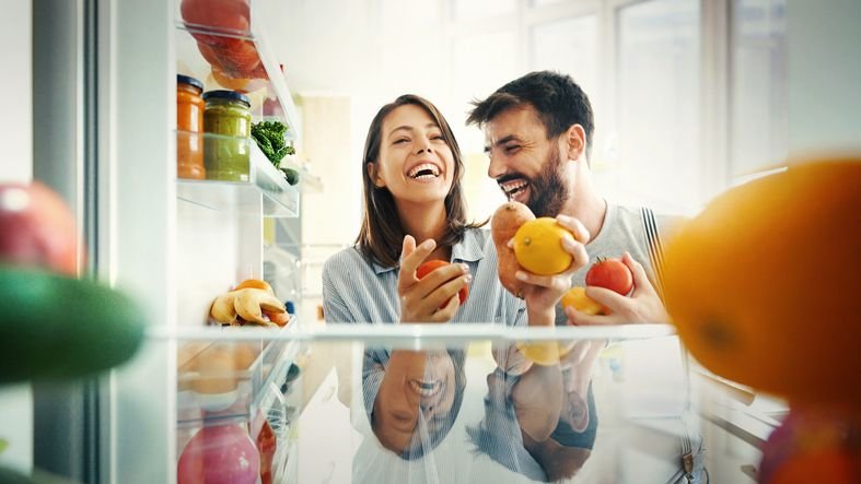 Casal sorridente em ângulo de uma foto batida de dentro de uma geladeira side by side em uma cozinha moderna, satisfeitos por terem escolhido a geladeira certa.