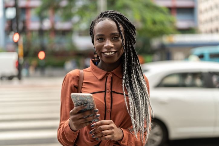 Mulher sorridente com tranças utilizando smartphone em rua movimentada, com ambiente urbano ao fundo.