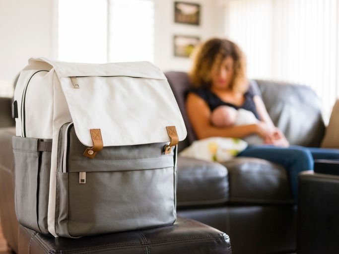 Mochila de bebê em destaque com mãe e recém nascido amamentando ao fundo.
