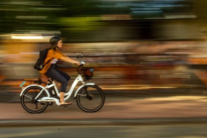 Mulher jovem e usando capacete preto, pedalando uma bicicleta elétrica branca em um trajeto urbano.