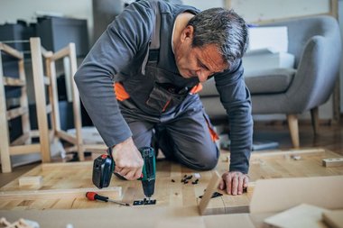 Homem usando furadeira parafusadeira pra perfurar uma peça de madeira na sua sala, em uma atividade comum de montagem.