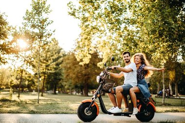 Duas pessoas andando em scooter elétrica em área urbana arborizada durante o dia