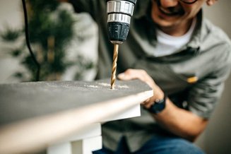 Homem usando furadeira elétrica pra perfurar uma peça de madeira, com foco na broca em ação durante um trabalho de montagem.