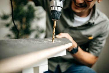 Homem usando furadeira elétrica pra perfurar uma peça de madeira, com foco na broca em ação durante um trabalho de montagem.
