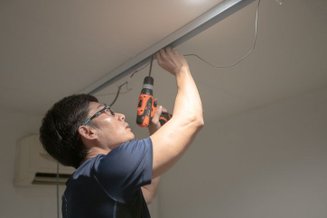 Homem segurando firmemente uma parafusadeira a bateria laranja e preta, parafusando um trilho de luz