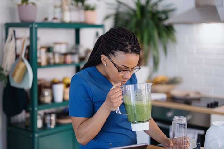 Mulher segurando um liquidificador com bebida verde na cozinha, observando o conte&uacute;do ap&oacute;s o preparo e lidando com a limpeza do aparelho de forma pr&aacute;tica e r&aacute;pida.
