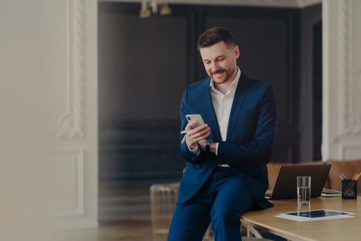Homem sentado na mesa de um escritório usando um terno azul escuro, sorrindo enquanto mexe no celular