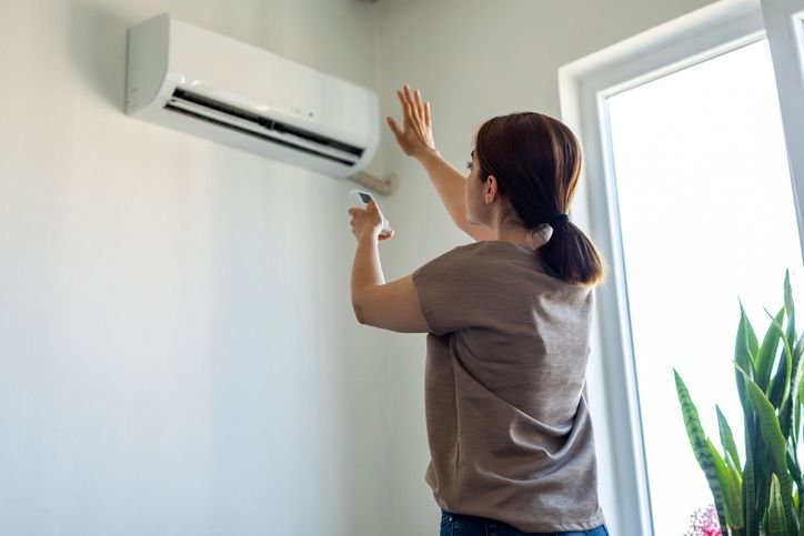 Mulher ajustando o ar-condicionado Philco em sua parede na sala de estar, com controle remoto na m&atilde;o, ambiente arejado e iluminado