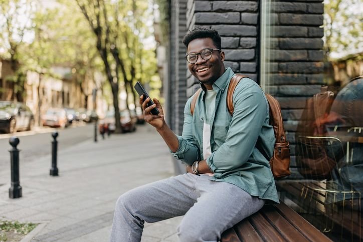 Homem com mochila masculina sentado em banco na rua, sorrindo e usando &oacute;culos enquanto verifica o celular, ambiente urbano com &aacute;rvores e carros ao fundo. 