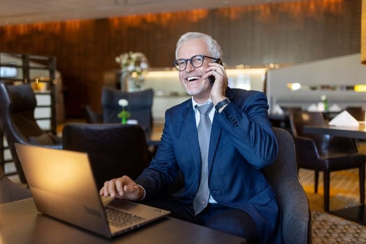 Homem num ambiente de trabalho usando terno enquanto fala no celular com um notebook na mesa