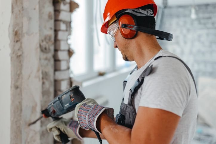 Profissional usando parafusadeira elétrica em obra, com capacete, luvas e protetor auricular, realizando trabalho de fixação em parede.