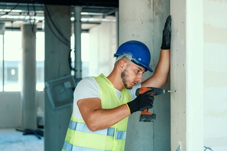 Profissional da constru&ccedil;&atilde;o civil usando parafusadeira el&eacute;trica em parede, com capacete, luvas e colete de seguran&ccedil;a, realizando instala&ccedil;&atilde;o em ambiente de obra.