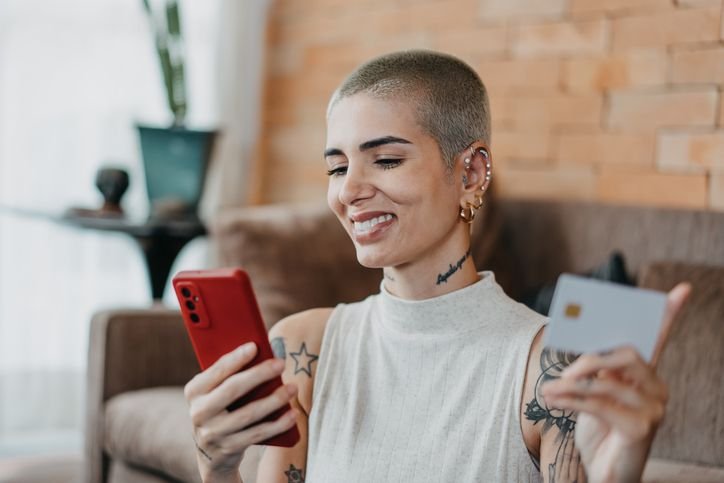 Mulher sorrindo com o celular em uma m&atilde;o e o cart&atilde;o de cr&eacute;dito na outra.