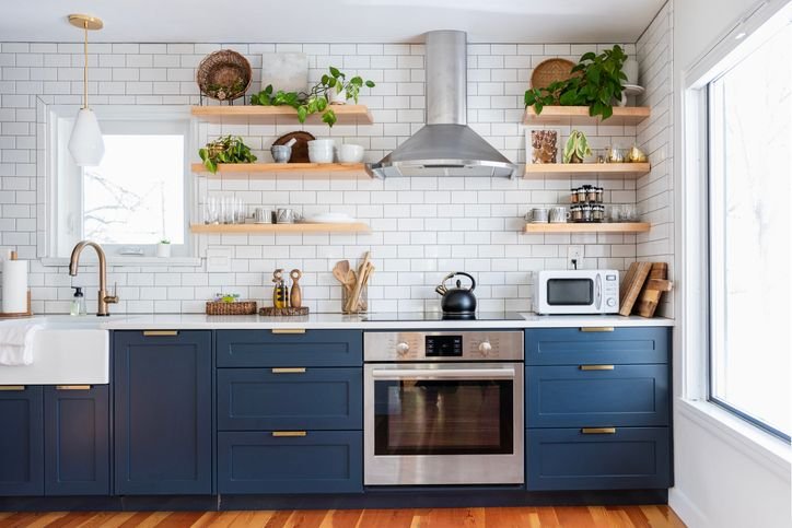 Cozinha moderna com arm&aacute;rios inferiores azul-marinho e puxadores dourados, bancada branca, pia com torneira dourada, fog&atilde;o e coifa de inox ao centro, backsplash de azulejos brancos tipo metr&ocirc; e prateleiras de madeira com plantas, lou&ccedil;as e objetos decorativos.