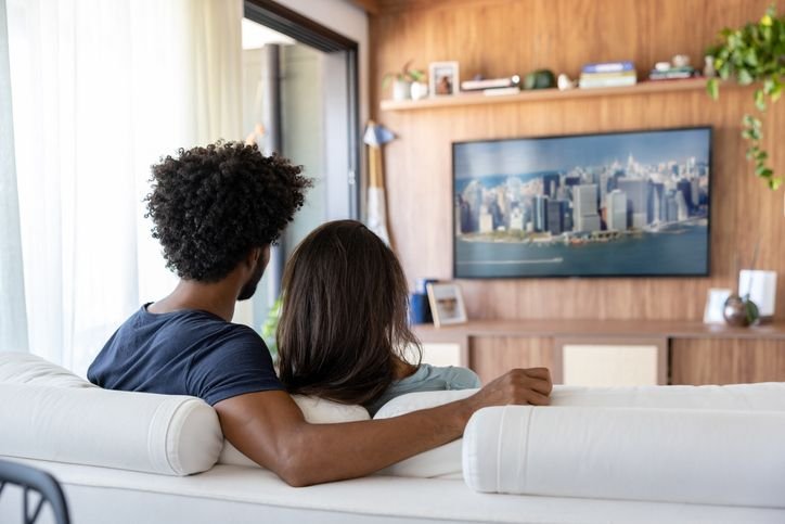Casal assistindo filmes em uma TV QLED 65 na sala de casa, aproveitando uma experiência mais imersiva e confortável. A tela grande com imagem vibrante transforma o ambiente em um verdadeiro cinema particular.
