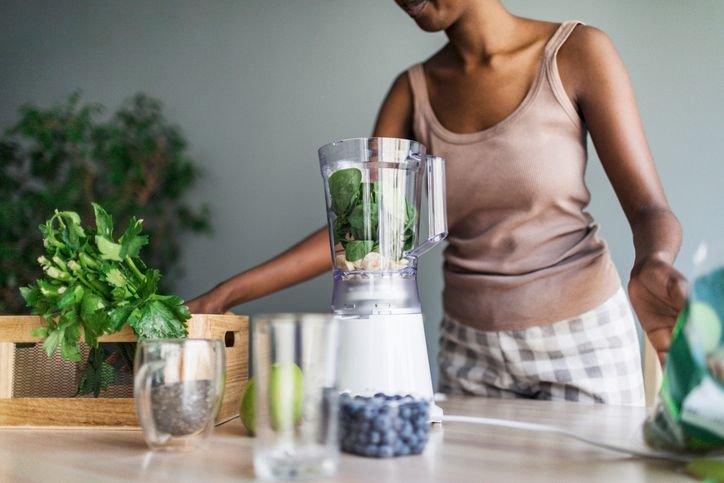 Mulher preparando ingredientes verdes em um liquidificador na cozinha, com atenção à higienização do aparelho após o uso para limpeza rápida e sem cheiro.