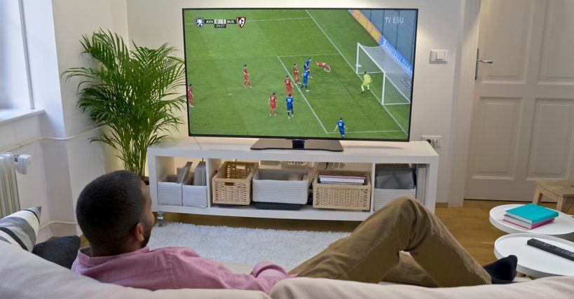 Homem assistindo jogo de futebol em uma Smart TV 85 polegadas, em sala de estar moderna. Imagem representa experiência de tela grande com alta imersão e qualidade 4K em ambiente doméstico.