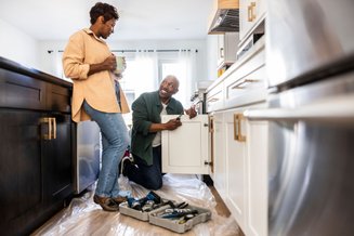 Imagem de casal montando armário de cozinha juntos; homem ajoelhado utiliza ferramentas enquanto a mulher observa segurando uma caneca, em ambiente claro e moderno.