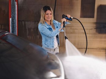 Mulher utilizando lavadora de alta pressão para limpar veículo, ajustando o jato de água com firmeza durante a higienização.