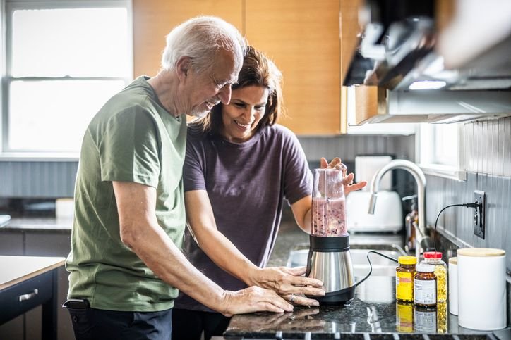 Casal sorridente na cozinha usando um liquidificador sobre a bancada, preparando uma bebida juntos enquanto ingredientes ficam ao lado.