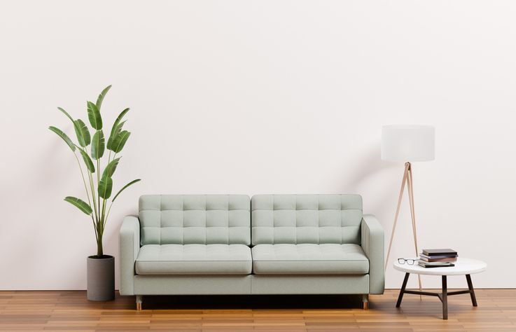 Sof&aacute; pequeno de cor cinza claro em sala moderna com parede branca, acompanhando uma planta decorativa, uma l&acirc;mpada de p&eacute; e uma mesa de canto com livros.