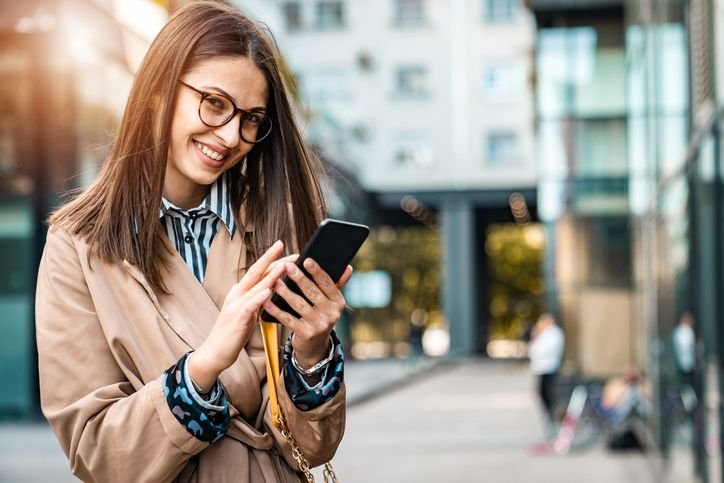 Mulher usando smartphone em ambiente urbano, com foco no uso do celular pra redes sociais, mensagens e conex&atilde;o 5G na rotina.