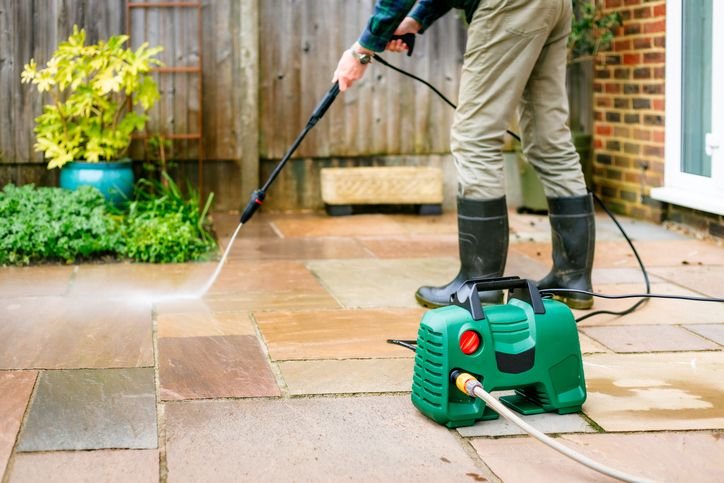 Pessoa usando uma lavadora de alta pressão portátil verde para limpar piso externo de pedra no quintal, com mangueira conectada e jato de água em ação.