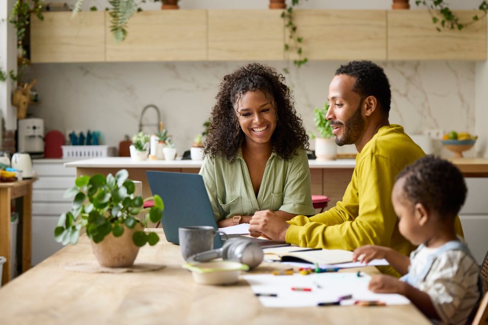 Casal olhando sorridente pra tela de um notebook, enquanto uma criança desenha na mesa ao lado deles. 
