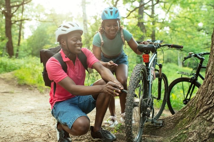 Duas pessoas usando capacetes de ciclismo param em uma trilha arborizada enquanto uma delas utiliza uma bomba de encher pneu pra ajustar a press&atilde;o da roda dianteira da bicicleta, sorrindo durante a atividade ao ar livre.