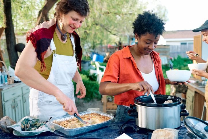 Duas mulheres em uma confraterniza&ccedil;&atilde;o ao ar livre, servindo comida em uma mesa com panelas el&eacute;tricas grandes e travessas, com vegeta&ccedil;&atilde;o ao fundo.