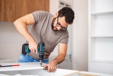Homem instalando armário de cozinha moderno utilizando uma parafusadeira durante o processo de montagem.