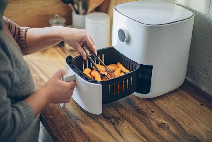 Pessoa retirando a cesta da air fryer enquanto prepara alimentos, mostrando o uso pr&aacute;tico da air fryer no dia a dia com preparo de receitas r&aacute;pidas e com menos &oacute;leo.  