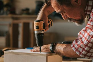 Homem mais velho usando uma parafusadeira sem fio pra fixar um parafuso em uma peça de madeira.